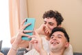 Snuggling gay couple in pajama laughing and using the smart phone. Cohabitation of homosexual couple