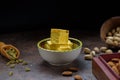 Soan papdi dessert cubes in bowl, cardamom grains in wooden scoop, pistachios and almond on concrete kitchen surface with black