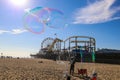 A soap bubble in the sky with people relaxing on the beach and a colorful Ferris wheel and rollercoaster on the pier