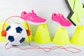 Soccer ball with red headphones, amid a row of yellow cones and pink sneakers, on a gray background