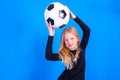 Soccer fans. Cute blonde girl in black shirt raised the ball over her head on blue studio background. Copyspace Royalty Free Stock Photo