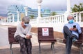 Social distancing concept. Two senior people sitting outdoor on allowed bench, using mobile phone