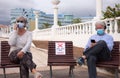 Social distancing concept. Two senior people sitting outdoor on allowed bench, using mobile phone