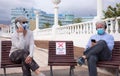 Social distancing concept. Two senior people sitting outdoor on allowed bench, using mobile phone