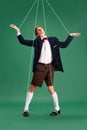 Social manipulation and pressure. Young guy, marionette on string against green studio background