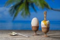 Soft boiled egg in eggcup with slice of toasted toast on wooden table with sea water, coconut palm tree and blue sky background on Royalty Free Stock Photo