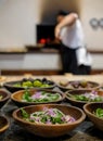 In soft focus in the background, chef puts pizza in pizza oven. In the foreground in focus, bowls of fresh salad.