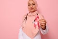 Focus on Breast Cancer Awareness Ribbon, in hands of blurred Muslim woman doctor in pink hijab, isolated on pink background, copy