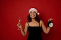 Soft focus on a champagne flute with sparkling wine and alarm clock in the hands of pretty woman in Santa hat smiling with