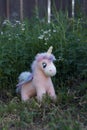 A soft pink toy unicorn stands on green grass against the background of a fence.