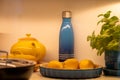 Softly lit kitchen scene with a yellow teapot, lemons dish, and a sleek blue bottle, creating a serene, domestic still