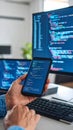 Software developer coding on a smartphone while working at a desk with multiple screens displaying program code Royalty Free Stock Photo