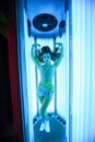 Solarium. Pretty girl with african braids in a dress for oriental dances sunbathing in a vertical sunbed. Blue neon glow