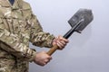 A soldier in an American military uniform holds a sapper shovel in his hands on a light background.