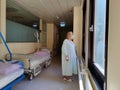 Solitary female patient wearing mask and hospital robe looking out trough big windows of hospital hallway.