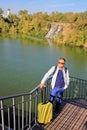 Solo traveling man with backpack and yellow luggage on the wooden bridge with church and waterfall background