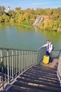 Solo traveling man with backpack and yellow luggage on the wooden bridge with church and waterfall background