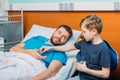 Son giving pills and glass of water to his sick father laying at ward