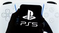 Sony PS5 logo seen on smartphone and official PS5 DualSense controller on the blurred screen at the background. Real photo, not a