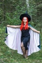 The sorceress in a hat and a black cloak in the forest divides over the sorceress with red hair in a hat and a black