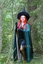 A sorceress with red hair in a pointed hat and a black cloak in the forest is engaged in charms with a candle. Royalty Free Stock Photo