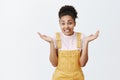 Sorry, oops. Portrait of cute attractive dark-skinned female in yellow overalls, shrugging with raised palms, smiling