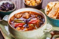 Soup with meat in bowl with oregano, chickpeas, peppers and vegetables served with crackers and bread on dark wooden background,