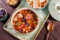 Soup with meat in bowl with oregano, chickpeas, peppers and vegetables served with crackers and bread on dark wooden background