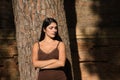 South American woman, young and beautiful, brunette, in brown dress, leaning on the trunk of a tree with her arms crossed, angry.