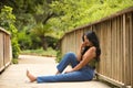 South American woman, young, beautiful, brunette, with colorful top and jeans sitting on a wooden bridge, pensive and with a lost