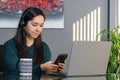 South Asian woman having a business call in headphones at a desk, typing on a phone in home office