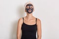 Spa teen girl applying black facial clay mask. Beauty treatments at home. Lady in sleeveless t shirt and hair band looking Royalty Free Stock Photo