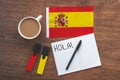 Spain national flag, notebook to write foreign words, word hello in spanish, two markers, cup of coffee, pen on brown wooden