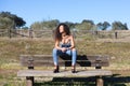 Spanish, brunette, curly-haired woman sits on a bench on the path leading to the forest. She is dressed in jeans and a white top Royalty Free Stock Photo