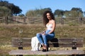 Spanish, brunette, curly-haired woman sits on a bench on the path leading to the forest. She is dressed in jeans and a white top Royalty Free Stock Photo