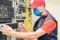 A specialist in a medical mask maintains equipment in the server room. A man in a red cap works in a datacenter. A technician