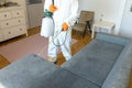 Person in protective suit with decontamination sprayer bottle disinfecting household and furniture