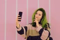 Spectacular green haired young attractive woman making selfie photo on smartphone, wears colorful sweater & toches her face