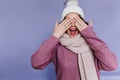 Spectacular woman in trendy knitted scarf covering eyes with hands. Indoor photo of laughing winsome woman in hat and