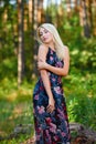 A spectacular young blonde woman with long hair elegantly poses outdoors against the background of trees in the summer.