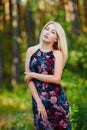 A spectacular young blonde woman with long hair elegantly poses outdoors against the background of trees in the summer.