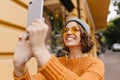 Spectacular young woman with happy smile making selfie with street on background. Cute girl wearing yellow sweater and