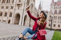 Spectacular young woman sitting on the ground with backpack and taking picture of herself. Outdoor shot of elegant