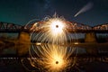 Spinning steel wool in abstract circle, firework showers of bright yellow sparks on long bridge reflected in river water under