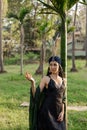 Spirited woman in long black off-the-shoulder dress stands leaning against palm tree on summer day.