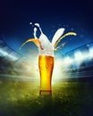 Splashes. Glass with lager foamy beer standing on grass at football stadium over evening sky with flashlights