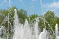 Splashing water from a fountain in the park against a background of blue sky and green trees. Refreshing water in the summer heat Royalty Free Stock Photo