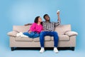 Split system. Young black couple with remote relaxing under air conditioner, sitting on couch over blue background Royalty Free Stock Photo