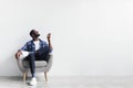 Split system. Young black man with remote turning on air conditioner, sitting in armchair against white wall, mockup Royalty Free Stock Photo