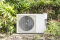 A split type inverter air conditioning condenser mounted outside a house partially concealed with shrubs. HVAC for tropical